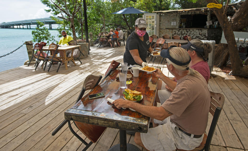 In this photo provided by the Florida Keys News Bureau, Alexeen Simms, a server at the Hungry Tarpon Restaurant in Islamorada, Fla., provides a luncheon entree to a couple Monday. June 1. 2020. After being closed to visitors since March 22, 2020, to help curtail the spread of COVID-19, the Florida Keys reopened to tourists Monday. Tourism employs about 45 percent of the Keys workforce. (Andy Newman/Florida Keys News Bureau via AP)