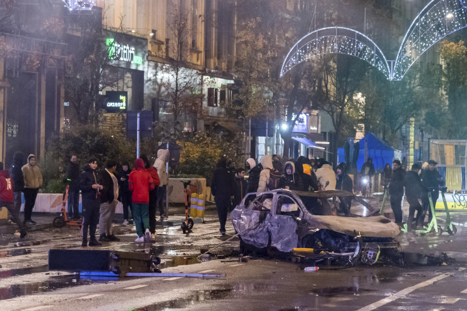 People stand next to a burned out car during riots in Brussels, Sunday, Nov. 27, 2022. Police had to seal off parts of the center of Brussels and moved in with water cannons and tear gas to disperse crowds following violence during and after Morocco's 2-0 win over Belgium at the World Cup. (AP Photo/Geert Vanden Wijngaert)