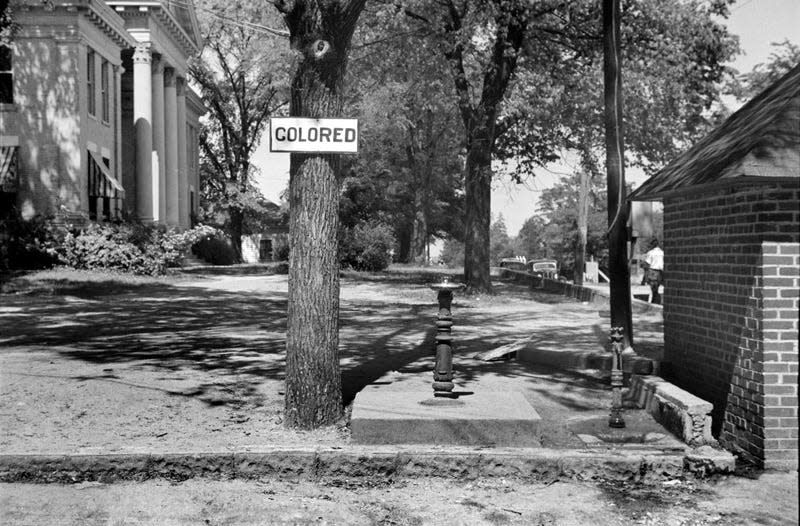 Drinking Fountain with Sign “Colored” nailed to Tree near County Courthouse, Halifax, North Carolina, USA, John Vachon, U.S. Farm Security Administration, April 1938. 