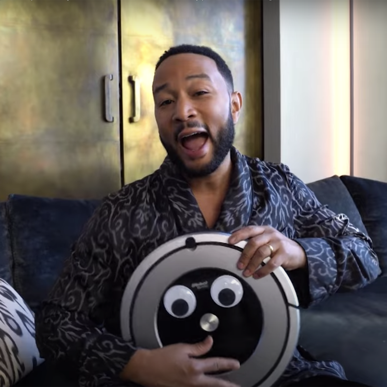 John Legend holds a roomba with googly eyes while sitting on a couch in a robe (The Tonight Show Starring Jimmy Fallon / NBC)