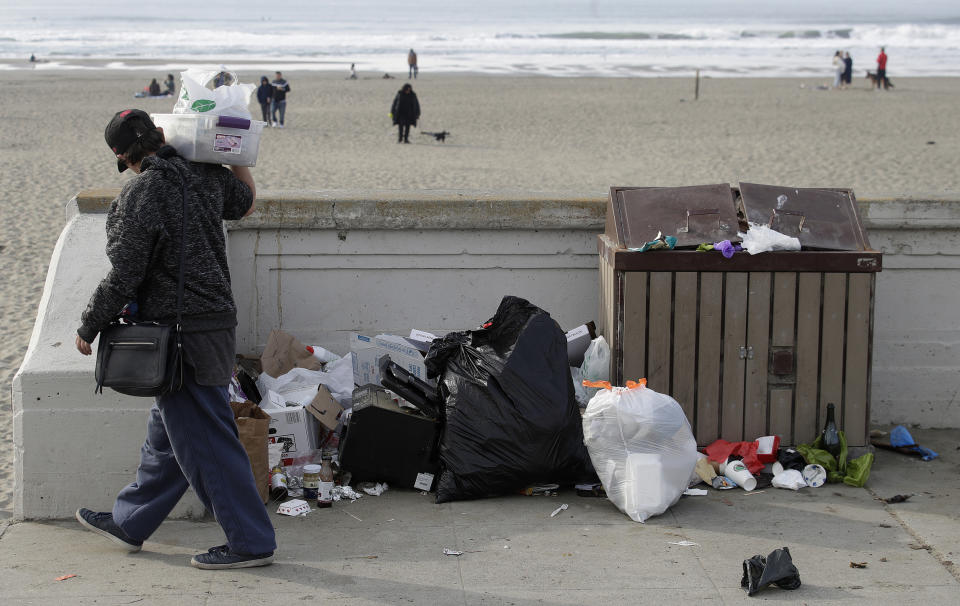 FILE - In this Jan. 3, 2019, file photo, a woman walks past trash piled next to a garbage bin at Ocean Beach in San Francisco. National parks across the United States are scrambling to clean up and repair damage caused by visitors and storms during the government shutdown while bracing for another possible closure ahead of the usually busy President's Day weekend. (AP Photo/Jeff Chiu, file)
