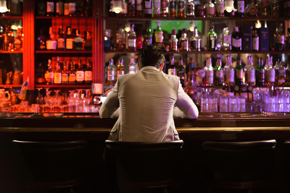 A man in a suit sits alone at a bar, looking at the rows of various liquor bottles lined up on the shelves behind the counter