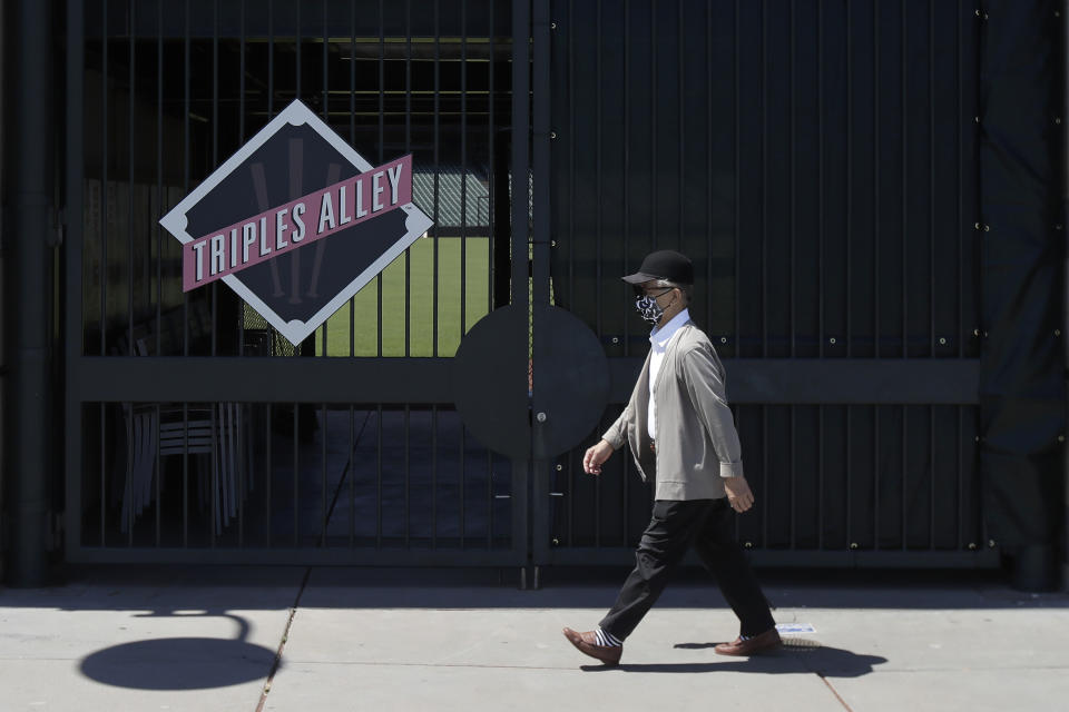 In his photo taken June 25, 2020, man wears a face mask during the coronavirus outbreak while walking past partially covered fencing outside Oracle Park, the San Francisco Giants' baseball ballpark, in San Francisco. This week, Major League Baseball players and owners reached an agreement to play an abbreviated, 60-game season that would start July 23 or 24 in teams’ home ballparks. But the seats will be empty. Instead, fans hoping to see a game in person will be have to settle for pressing their faces up against hotel windows, squinting through metal grates or climb to rooftops when baseball returns this month in otherwise empty stadiums. (AP Photo/Jeff Chiu)