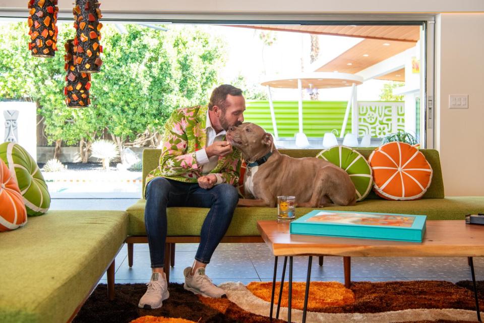 A man sits on a couch with his dog. The large window behind him looks out onto a yard.