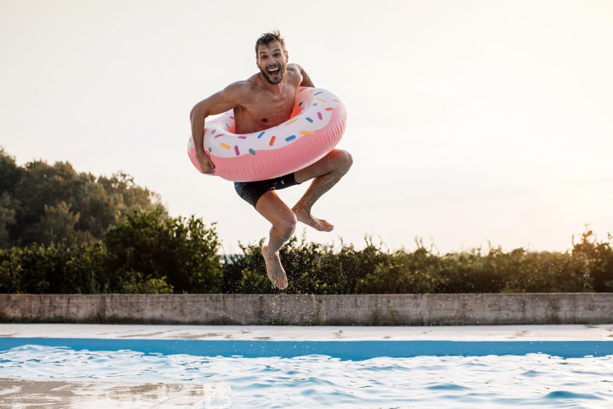 Happy young man in mid-air jumping with an inflatable ring into an in-ground swimming pool with edge of lawn and sun setting in background