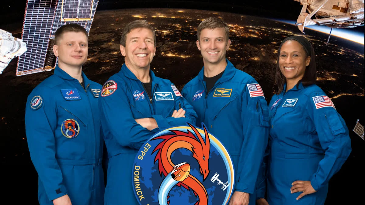  Three white men and a black woman stand smiling wearing blue astronaut flight suits. the circular graphic of the crew's mission patch, with an orange dragon spiralled into an 8, is in front of the group. a view of Earth at night, as seen from the space station is their backdrop. 