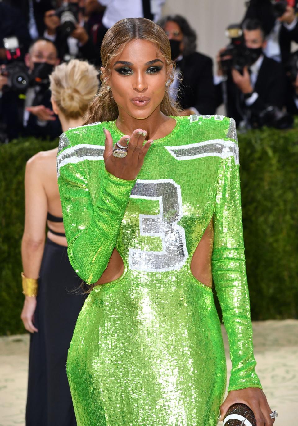 Ciara at an event blowing a kiss to the camera. She's wearing a football jersey-inspired sequined gown and her hair in a long ponytail