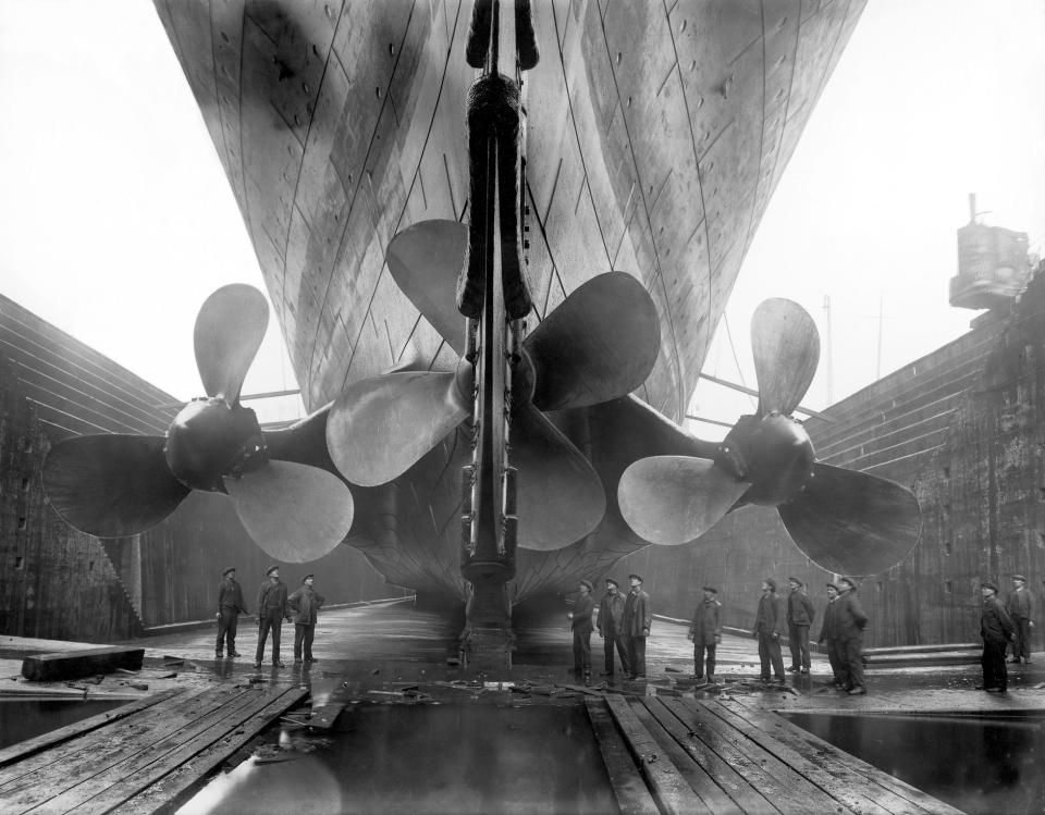 Workers stand in front of Titanic’s massive propellers in a shipyard. The impressive scale of the ship's construction is prominently displayed