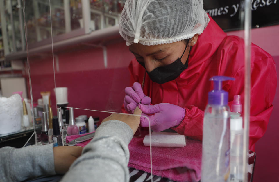 A manicurist paints a client's nails from behind a clear, plastic wall and wears a face mask, hat and gloves as a precaution amid the new coronavirus pandemic at a nail salon in La Paz, Bolivia, Friday, May 22, 2020. The nail salon re-opened three days ago and only attend clients with appointments in the mornings. (AP Photo/Juan Karita)