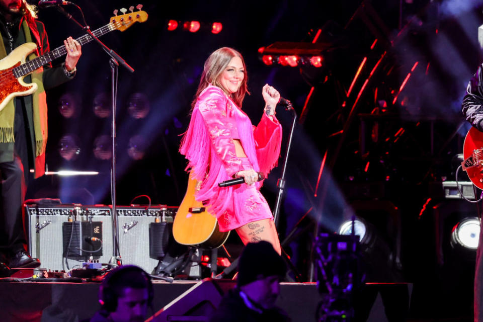 Elle in a fringed jacket performing on stage with a guitarist nearby