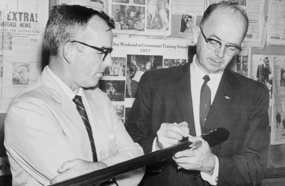 A white man dressed in a dark suit writes on a piece of wood as another white man watches.