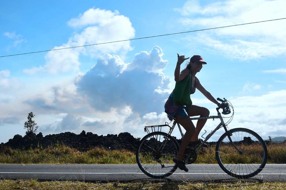 A cyclist offers the Shaka sign, also known as "Hang Loose" while riding past a plume of volcanis smoke in the distance over the area of Leilani Estates near the town of Pahoa