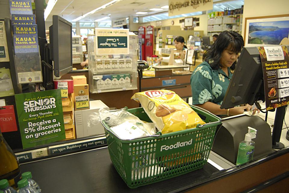 A worker scans items in a checkout line at a Foodland supermarket in Hawaii as a basket of bagged chips sits on the conveyor belt and maps of Hawaiian islands hang above the checkstand.