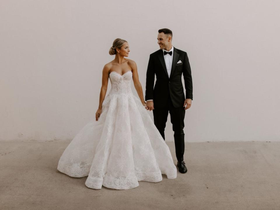 A bride and groom hold hands in front of a white wall.