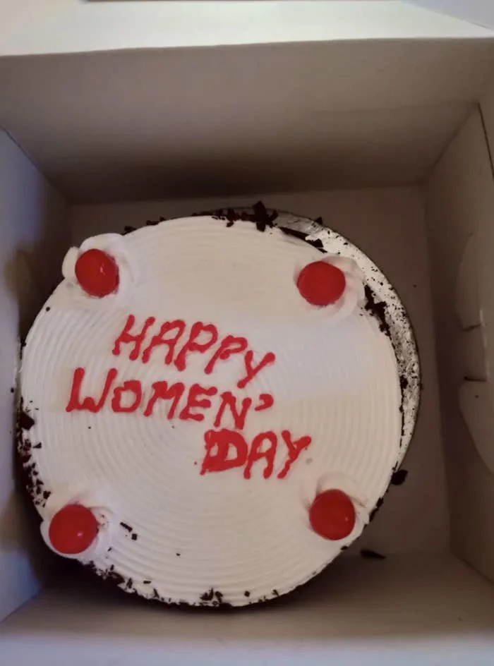 A cake with "HAPPY WOMEN' DAY" written on it erratically in icing, adorned with four cherries