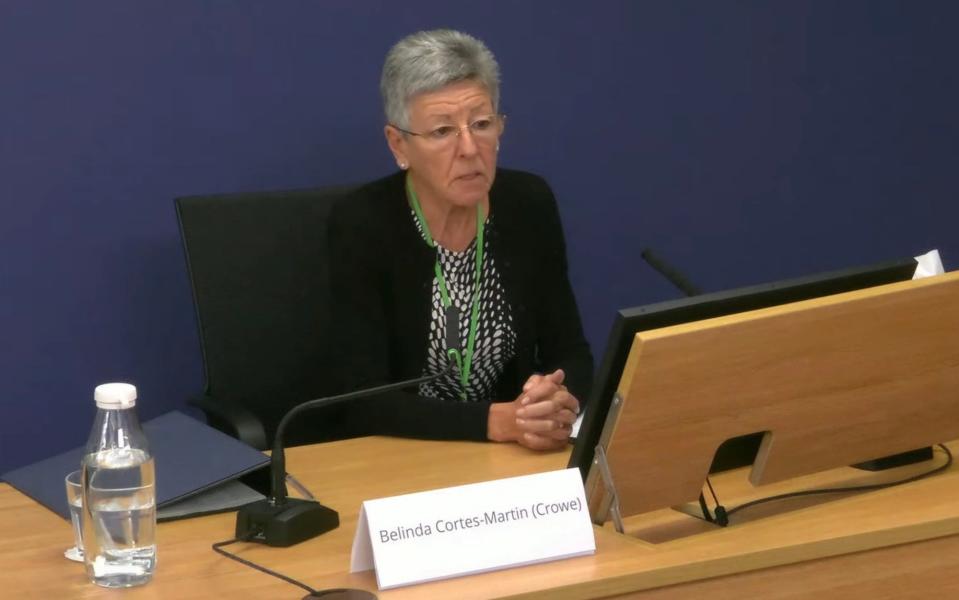 Mrs Cortes-Martin sits with hands folded at a desk to give evidence, with a microphone and a bottle of water