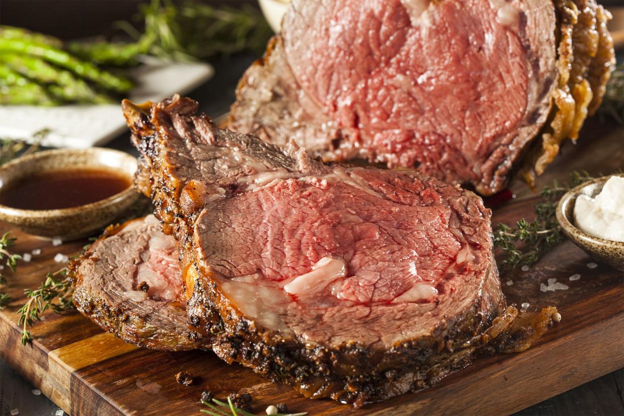 Closeup of cut slow-roasted beef prime rib with horseradish sauce on an wooden cutting board with dipping sauce and asparagus in the background