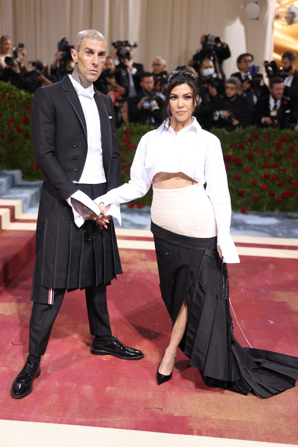 Travis Barker and Kourtney Kardashian attend the 2022 Met Gala. Travis wears a black suit with a skirt over his pants. Kourtney wears a cropped white button down and a skirt made of deconstructed meanswear.