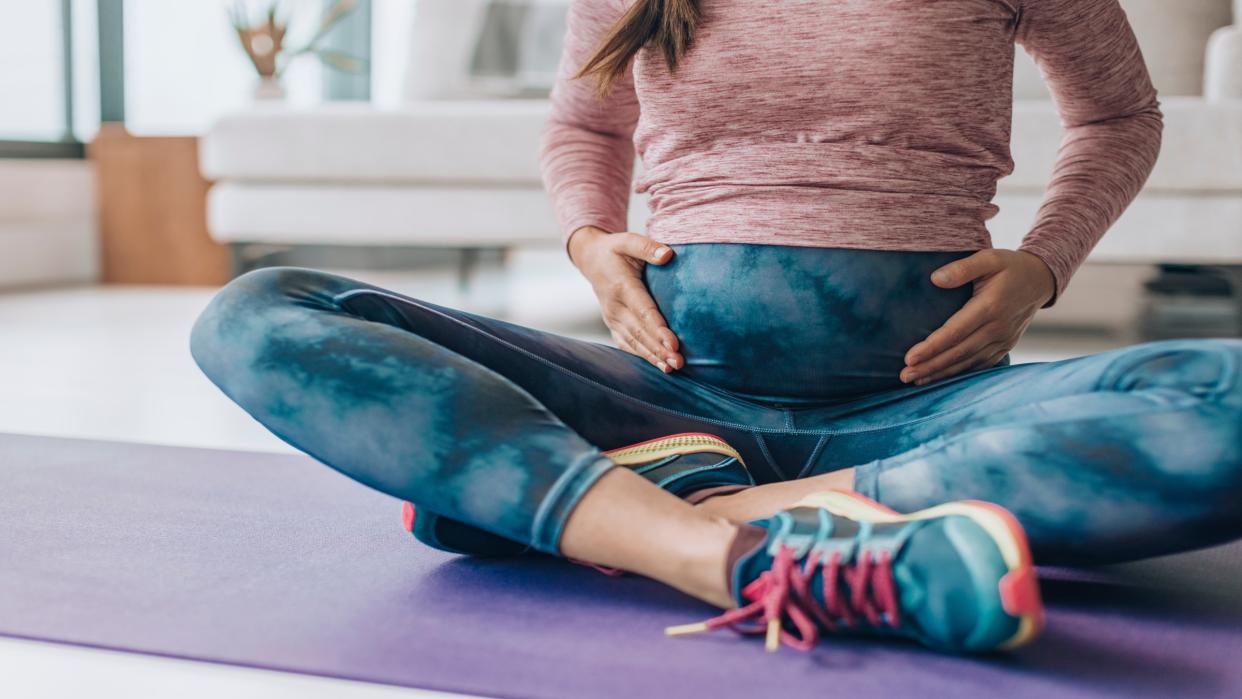  Pregnant woman shown in exercise clothes and sneakers, holding her stomach and sitting on an exercise mat in her home  