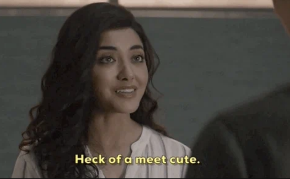 A woman with long, wavy hair and a light blouse is smiling and facing a person offscreen. Subtitle reads, "Heck of a meet cute."