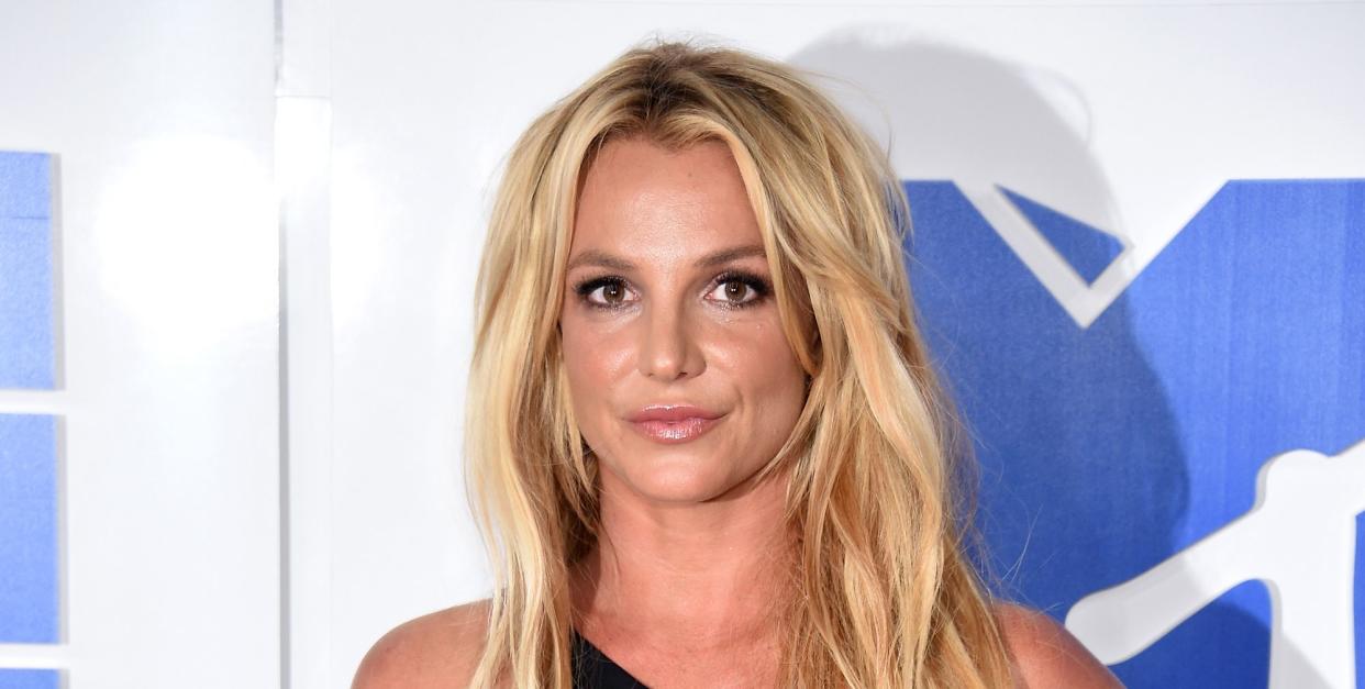 britney spears standing looking at the camera with left hand on hip, long blonde hair worn down in loose waves, wearing an asymmetrical long black dress with thigh split