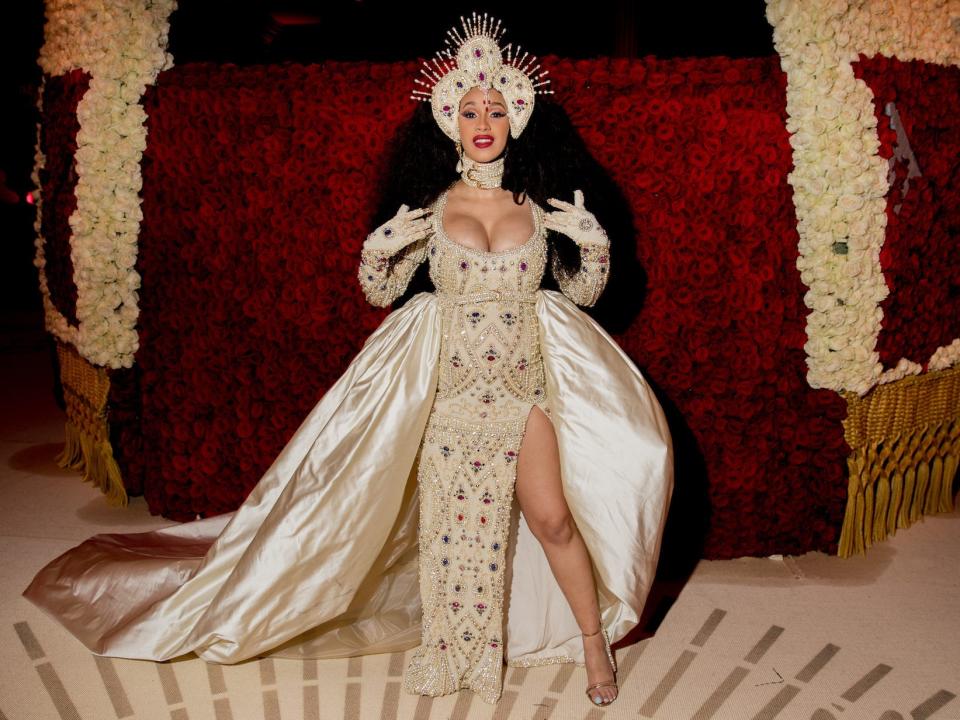 Cardi B poses in front of a wall of roses wearing a bejeweled gold look with an elaborate headpiece.