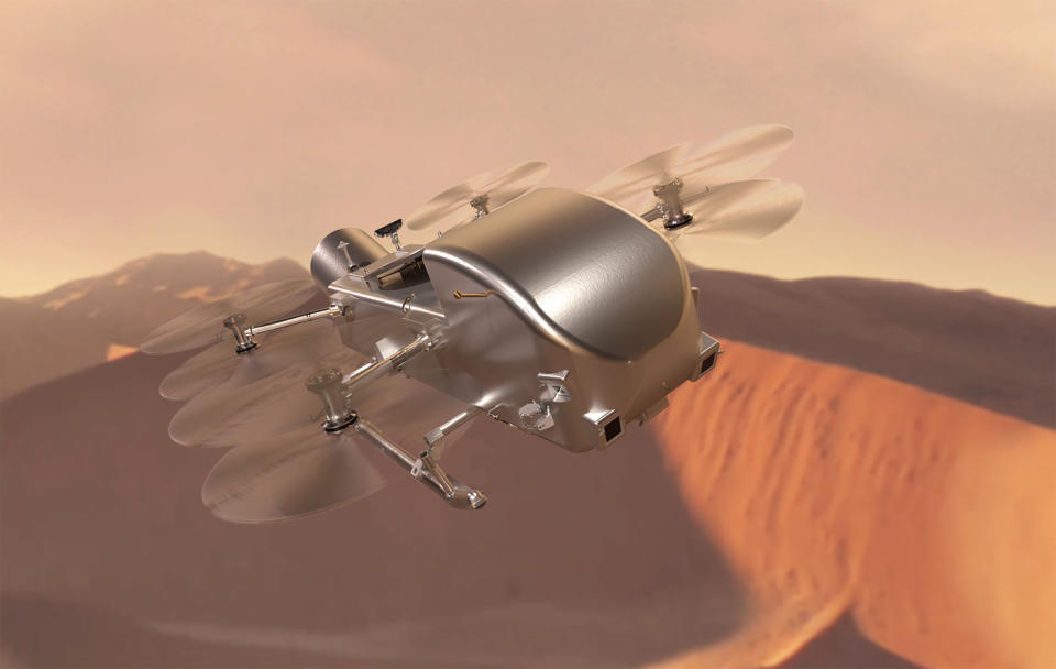Длинный серебристо-хромированный дрон с шестью пропеллерами на уровне тела напоминает стрекозу.  он летит над розово-коричневыми песчаными дюнами.