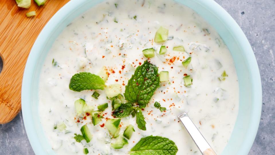 raita in a bowl with cucumbers, mint, and powdered red spice cutting board with mint and cucumbers alongside