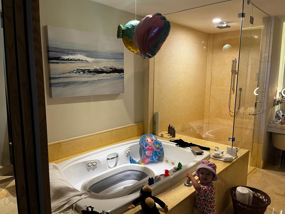 A bathtub in a hotel bathroom decorated with mermaid and seashell balloons. A small child stands in front of the tub