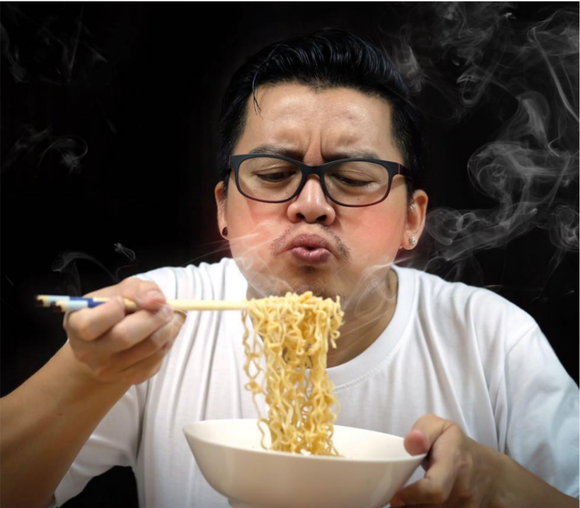 A young Chinese man with glasses eating a bowl of noodles with chop sticks.