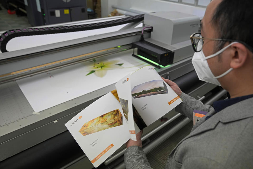 Wilson Tong, CEO of LifeArt Asia, looks at a catalogue showing over 40 different choices of coffin designs as a machine is printing the Lily picture on a cover of a coffin, at Tong's factory in Hong Kong, Friday, March 18, 2022. Hong Kong is running short of coffins during its deadliest outbreak of the coronavirus pandemic, which has cost about 6,000 lives so far this year. LifeArt, a company in Hong Kong is trying to make an alternative, cardboard coffin, which it says is environmentally-friendly. (AP Photo/Kin Cheung)