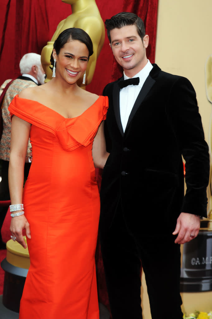 Paula Patton in an elegant off-shoulder gown and Robin Thicke in a black tuxedo pose together at an event with gold statues and red drapes in the background