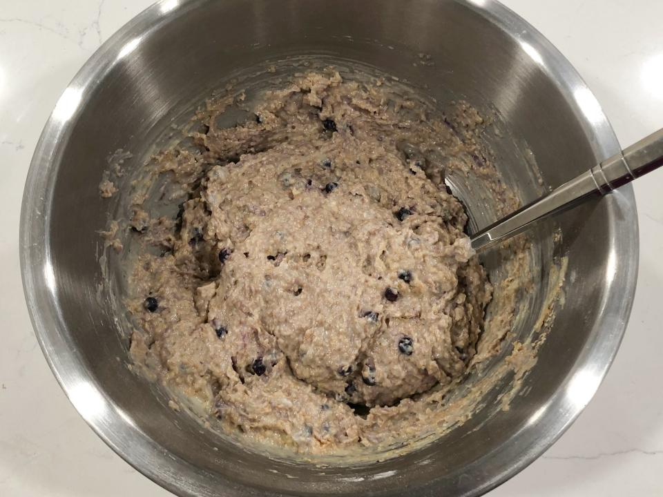 Muffin batter, which now includes blueberries and is a darker color, in a stainless-steel bowl with a spoon. 