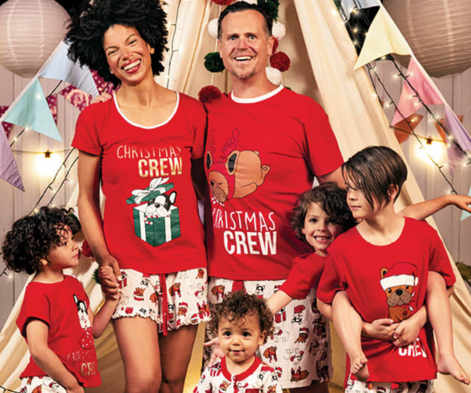 A family wear matching red Christmas pyjamas infront of a cream teepee with fairy lights and colourful bunting. 