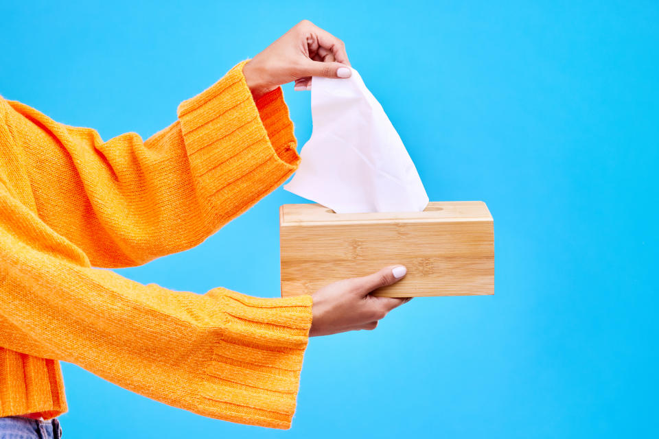 Person in an orange sweater taking a tissue from a wooden box against a blue background