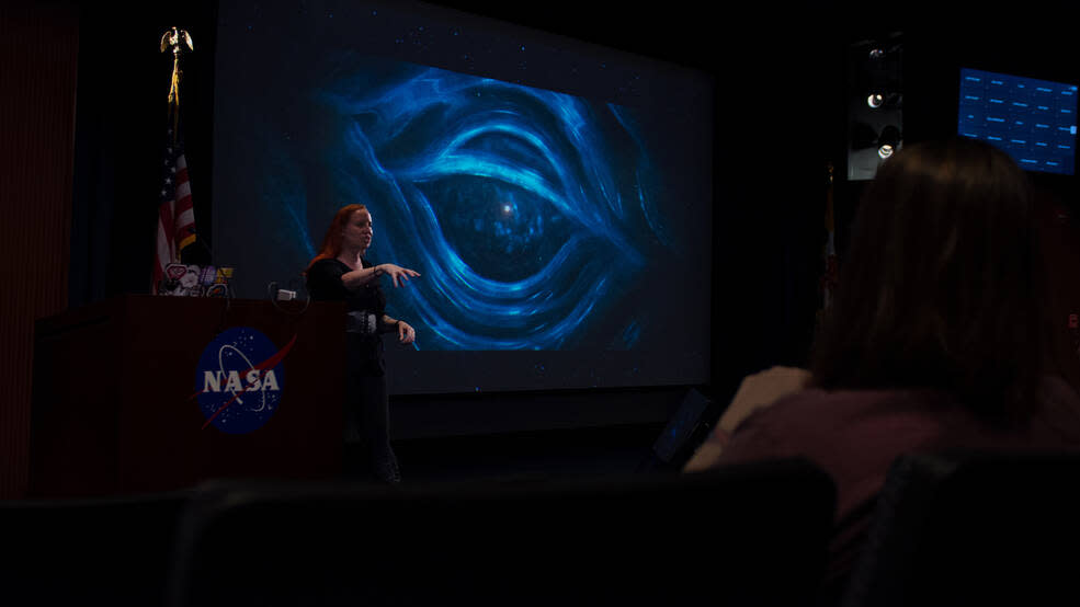  A woman gives a lecture in a dark room with space imagery projected on a screen in the background. 
