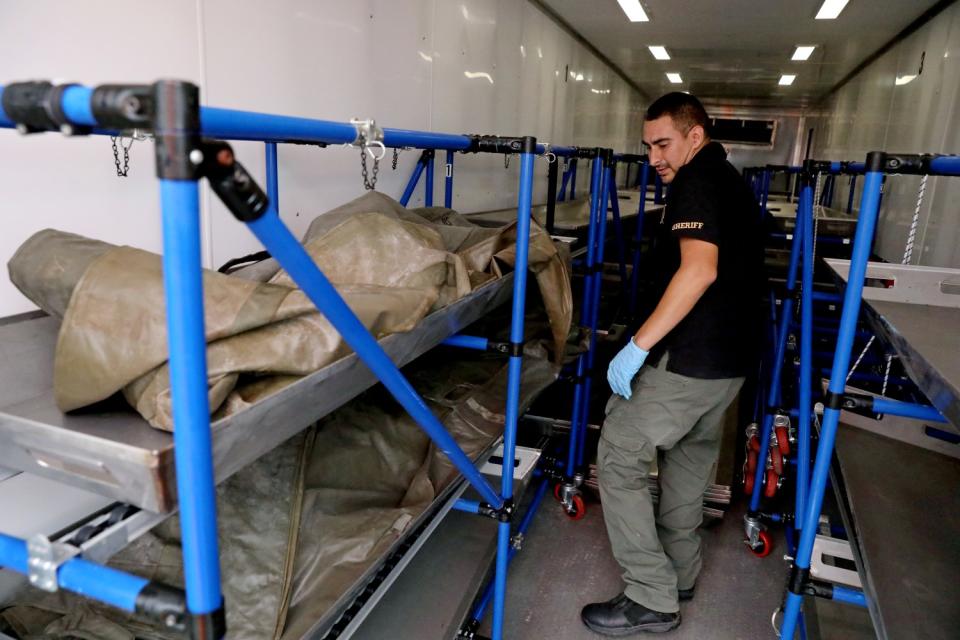 A man looks at a wrapped body on a rack.