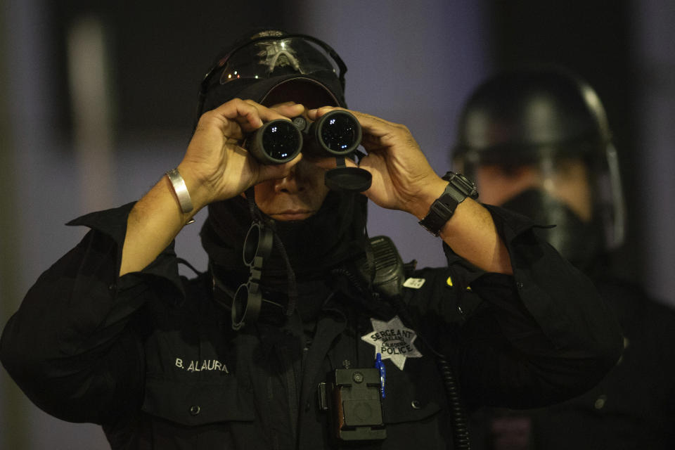 An Oakland police officer surveils protesters with binoculars during a protest on Saturday, July 25, 2020, in Oakland, Calif. Protesters in California set fire to a courthouse, damaged a police station and assaulted officers after a peaceful demonstration intensified late Saturday, Oakland police said. (AP Photo/Christian Monterrosa)