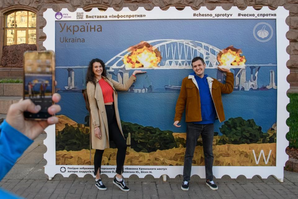 People pose for photos and take selfies in front of the large poster form of postage stamp depicting the Crimean Kerch Bridge on fire in Kyiv, Ukraine, October 08, 2022.