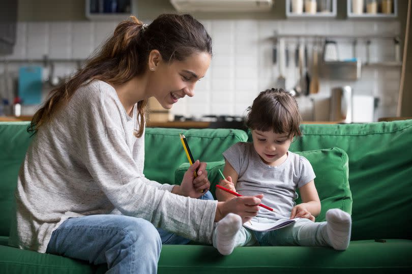 Smiling baby sitter and preschool kid girl drawing with colored pencils sitting on sofa together, single mother and child daughter playing having fun, creative family activities at home concept