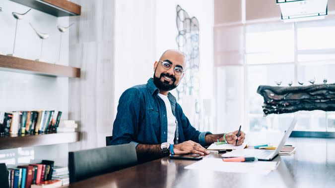 Cheerful handsome ethnic man with beard sitting with laptop and notepad at table in trendy office smiling away to colleague.