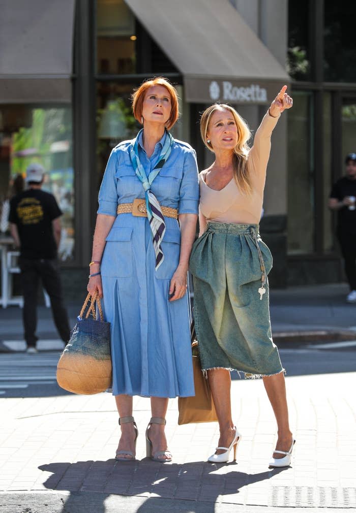 Cynthia Nixon wearing a blue dress and Kristin Davis in a beige top and green skirt, standing on a street. Kristin points while both look in the same direction