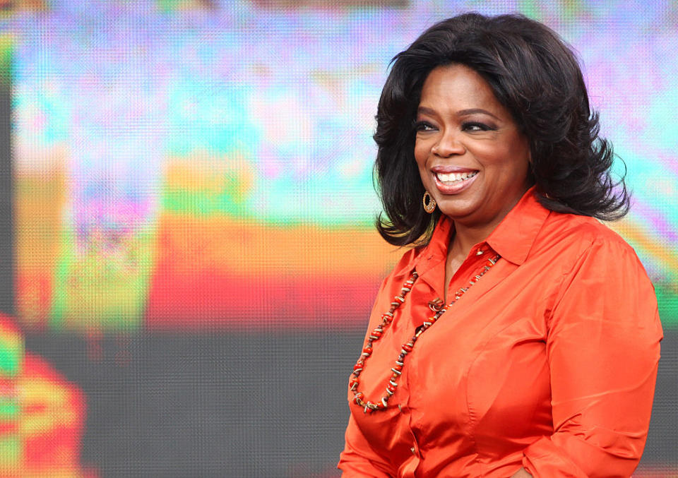 Oprah Winfrey in an orange blouse with a beaded necklace, smiling onstage