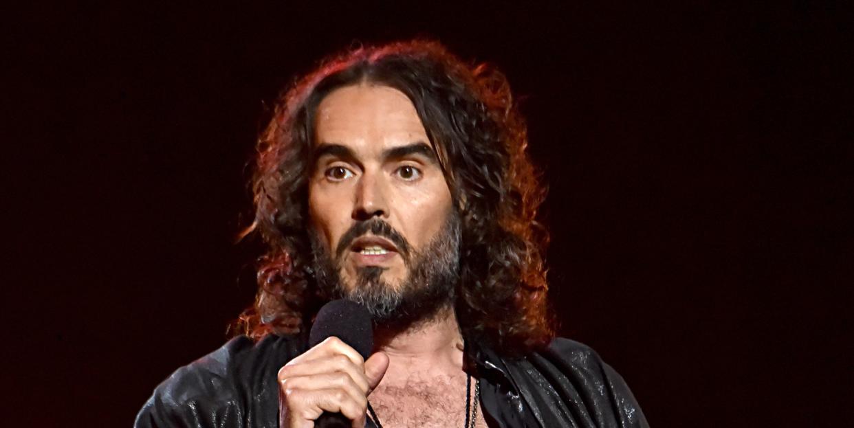 russell brand speaks at an event in 2020, he has long dark hair and a beard, is wearing an all black outfit and has a microphone in his hand