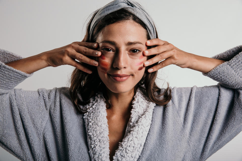 A woman wearing a cozy robe and headband applies skincare or makeup to her face, smiling gently at the camera. She has red stripes on her cheeks