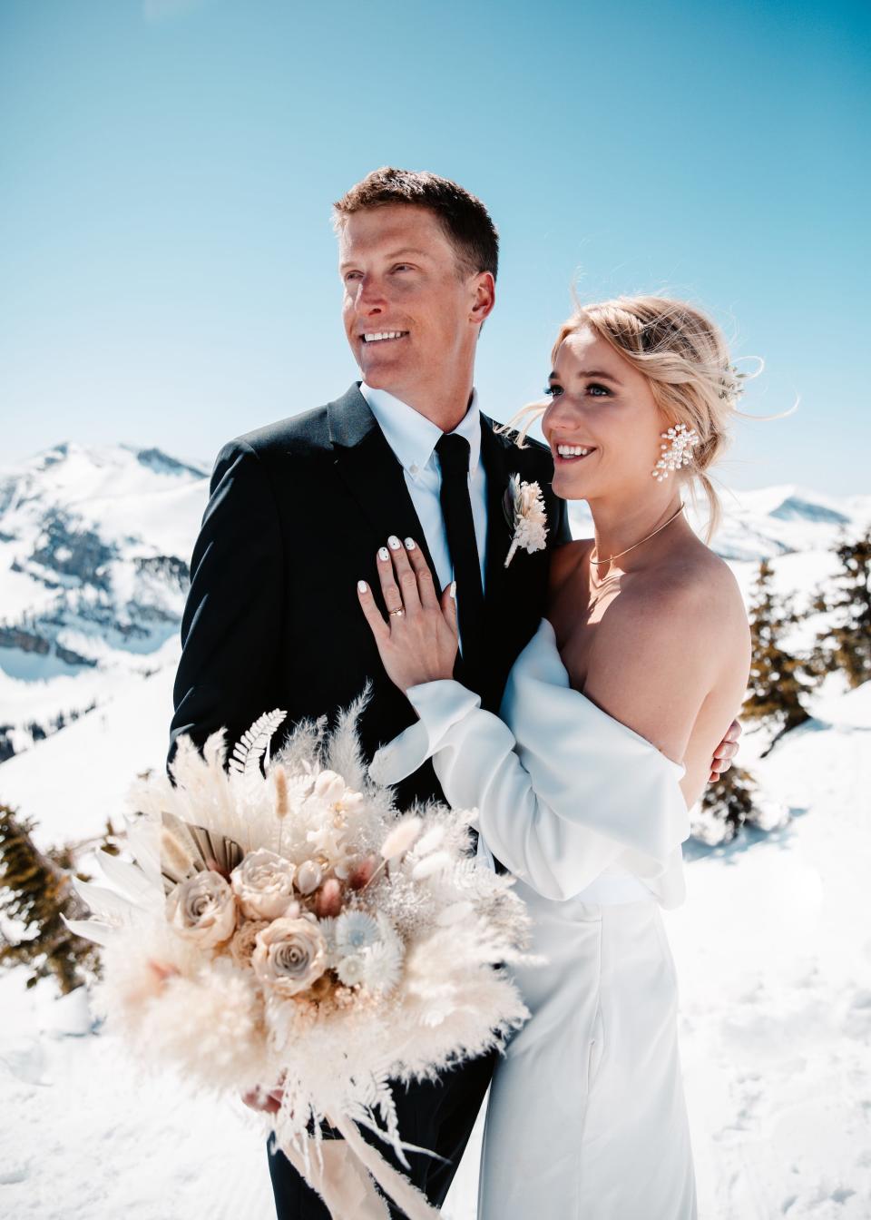 A bride and groom stand together on top of a mountain and look off into the distance.