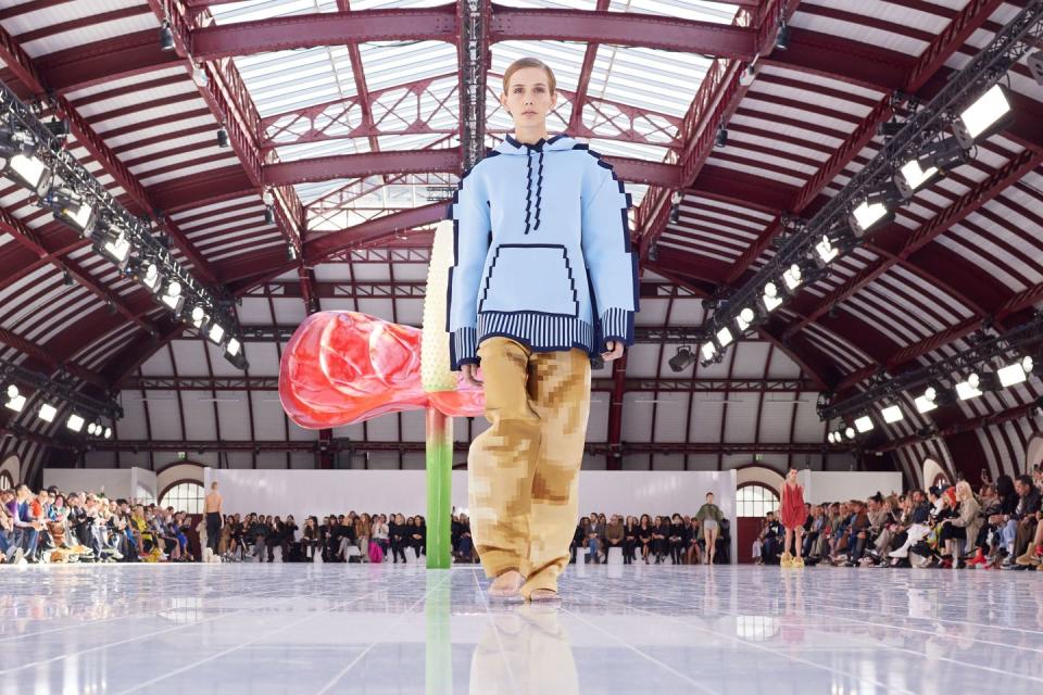 a blonde model in a blue shirt and yellow pants walks the runway at loewe spring 2023 in front of an oversized anthurium sculpture