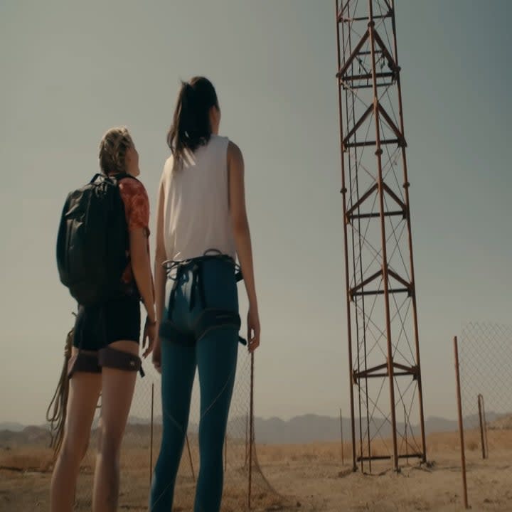 Two young women stand at the base of an extremely tall, rusty tower.