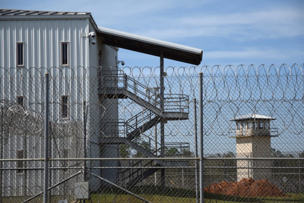 A three story prison dorm with aluminum siding and outside stairs rises behind a barbed wire fence. A guard tower can be seen in the background.