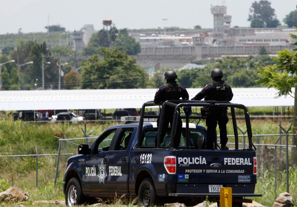 Two police are seen standing in the back of a vehicle and facing a prison complex, seen in the distance.